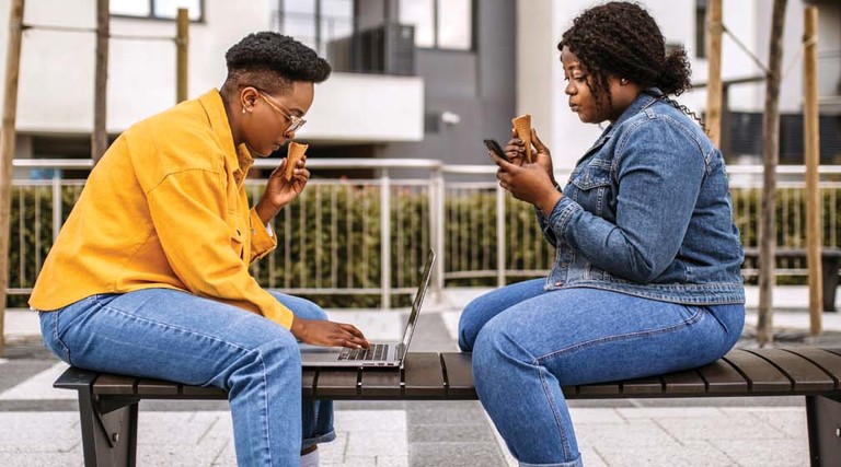 Two people sit on a bench while using their devices