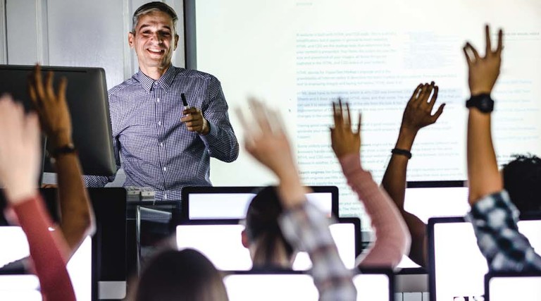 A professor stands at the front of a classroom as students raise their hands