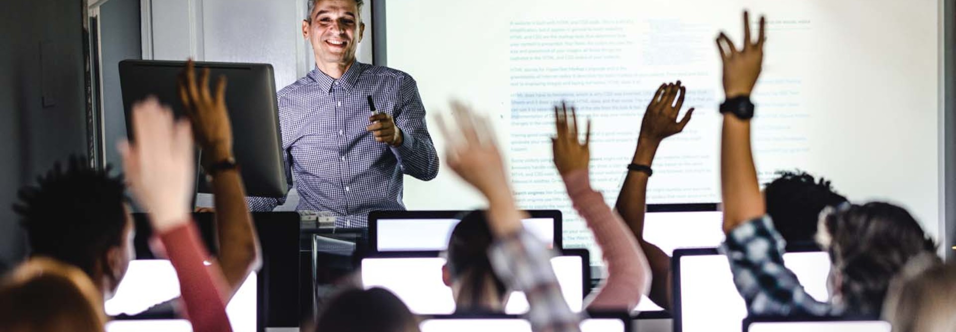 A professor stands at the front of a classroom as students raise their hands