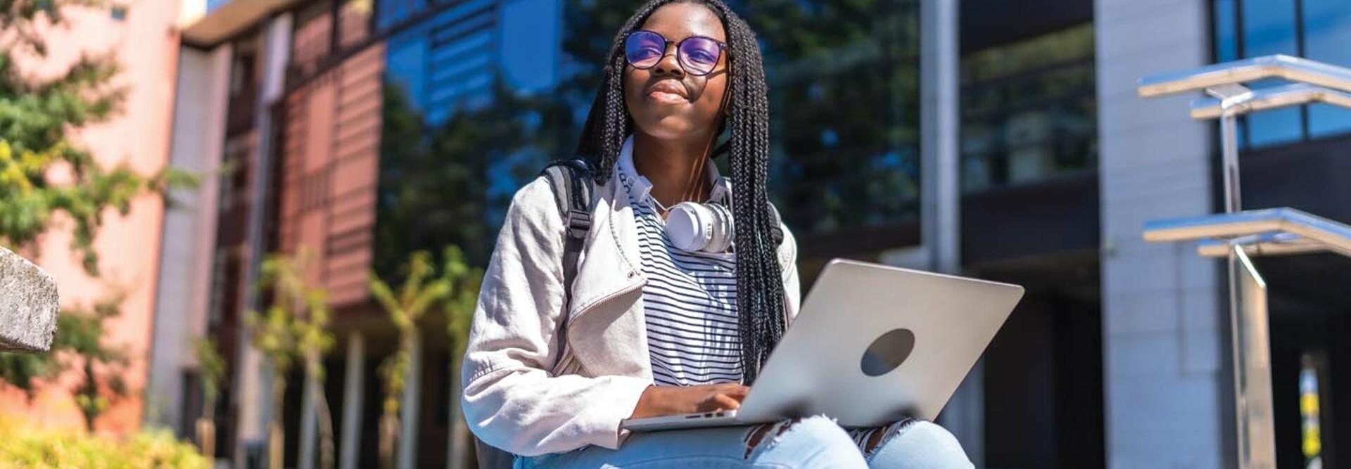 A college student with a laptop on her lap gazes skyward outside a campus building