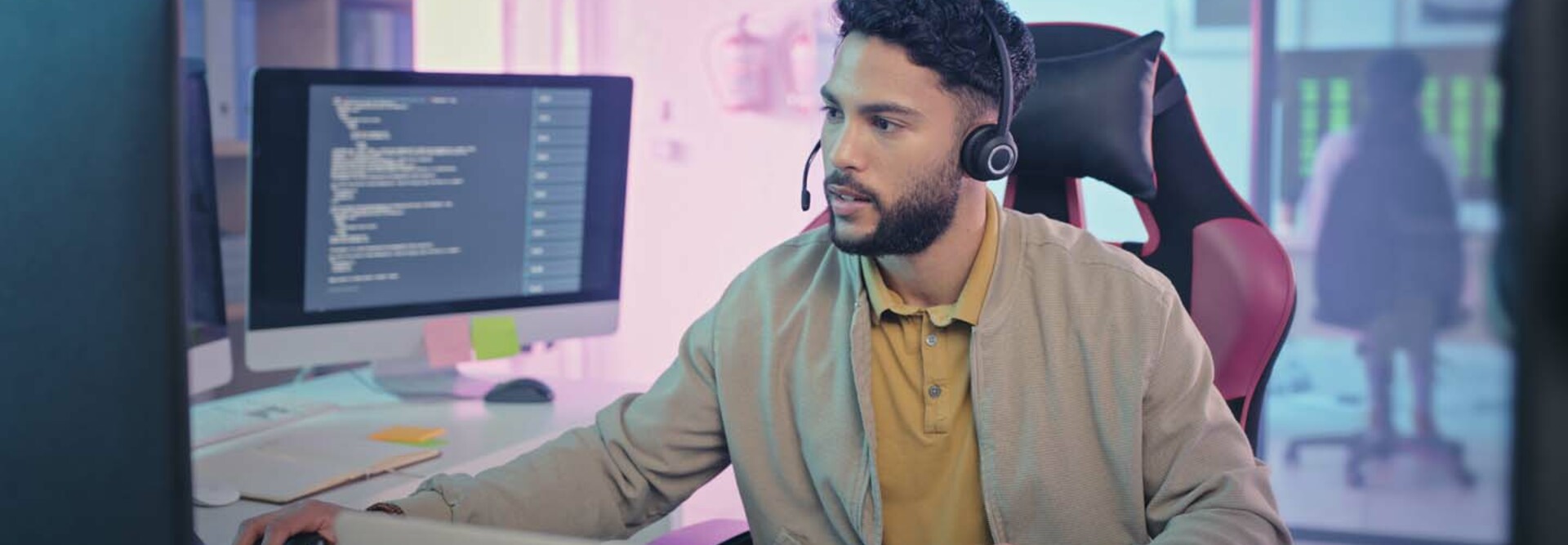 A man wearing a headset sits at a modern workstation with multiple monitors