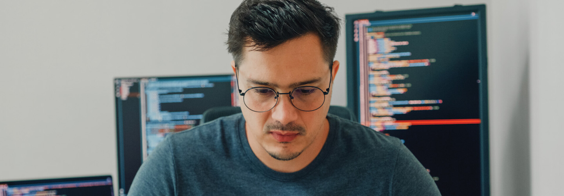 An unhappy looking man stares down intently, with a pair of computer screens displayed behind him
