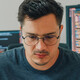 An unhappy looking man stares down intently, with a pair of computer screens displayed behind him