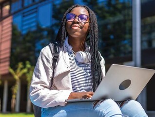 A college student with a laptop on her lap gazes skyward outside a campus building