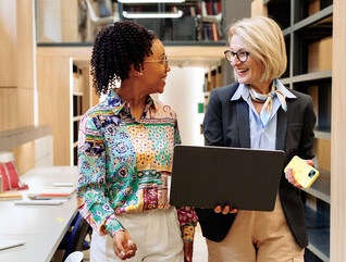 Two women stand and talk while one holds a laptop