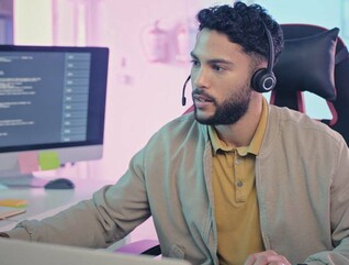 A man wearing a headset sits at a modern workstation with multiple monitors
