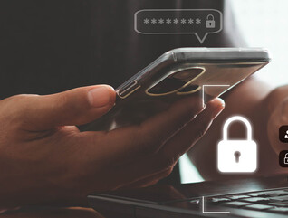 A person with a phone in one hand and their other hand on a keyboard, presumably to complete multifactor authentication