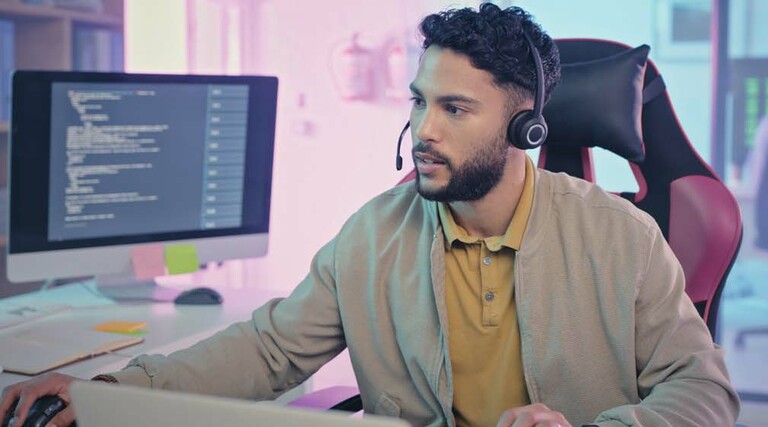 A man wearing a headset sits at a modern workstation with multiple monitors