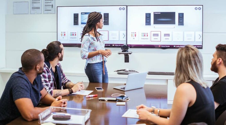A person stands at the front of a meeting room with a pair of monitors behind them.