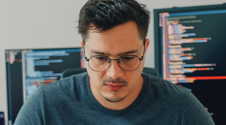 An unhappy looking man stares down intently, with a pair of computer screens displayed behind him