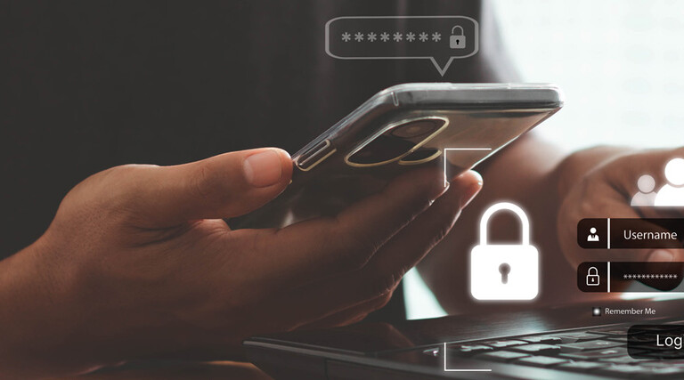 A person with a phone in one hand and their other hand on a keyboard, presumably to complete multifactor authentication