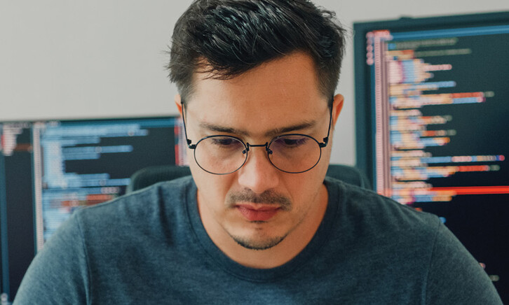 An unhappy looking man stares down intently, with a pair of computer screens displayed behind him