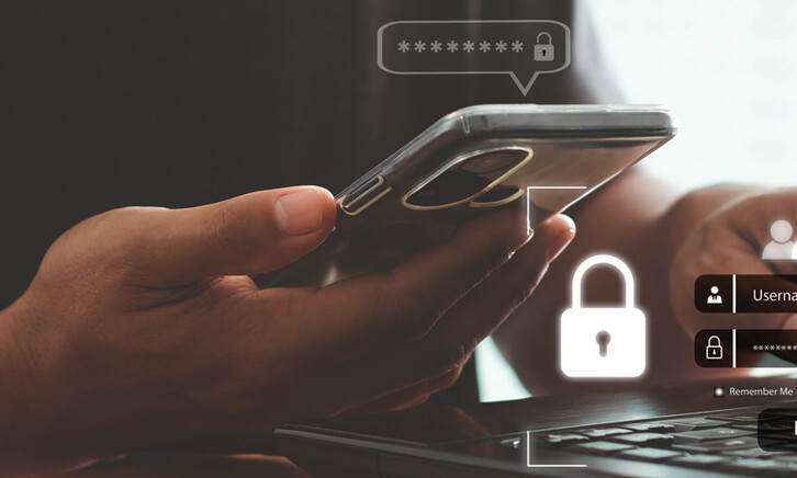 A person with a phone in one hand and their other hand on a keyboard, presumably to complete multifactor authentication