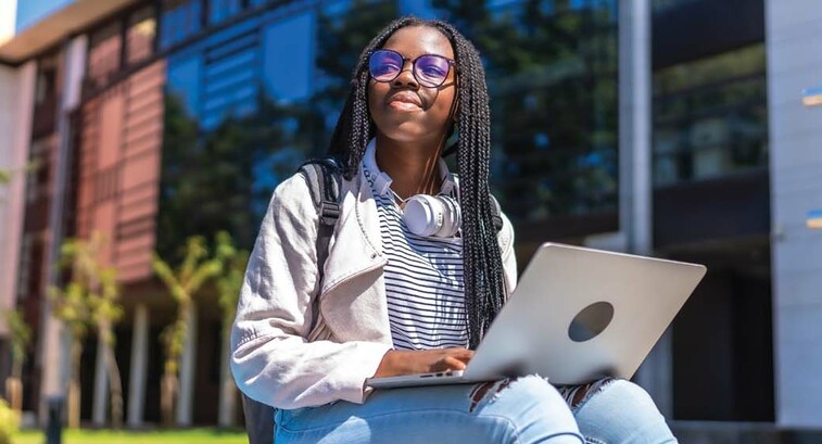 A college student with a laptop on her lap gazes skyward outside a campus building