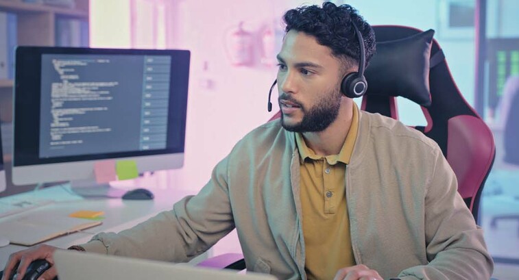 A man wearing a headset sits at a modern workstation with multiple monitors