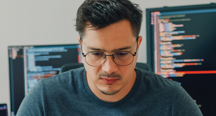 An unhappy looking man stares down intently, with a pair of computer screens displayed behind him