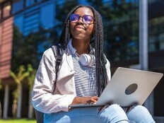 A college student with a laptop on her lap gazes skyward outside a campus building