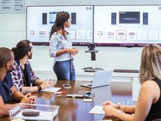 A person stands at the front of a meeting room with a pair of monitors behind them.