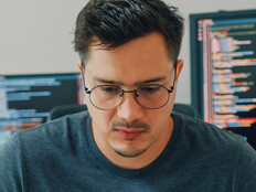 An unhappy looking man stares down intently, with a pair of computer screens displayed behind him
