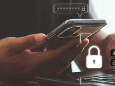 A person with a phone in one hand and their other hand on a keyboard, presumably to complete multifactor authentication