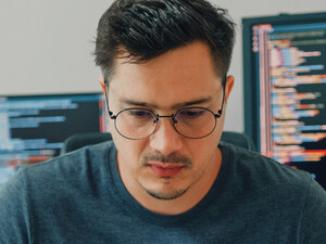 An unhappy looking man stares down intently, with a pair of computer screens displayed behind him