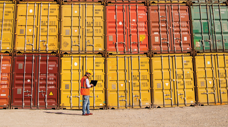 Supply Chain Disruptions - dock worker with laptop standing in front of cargo containers stacked two high