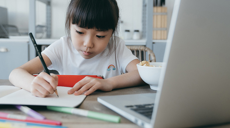 young girl taking notes on paper from computer while holding tablet 