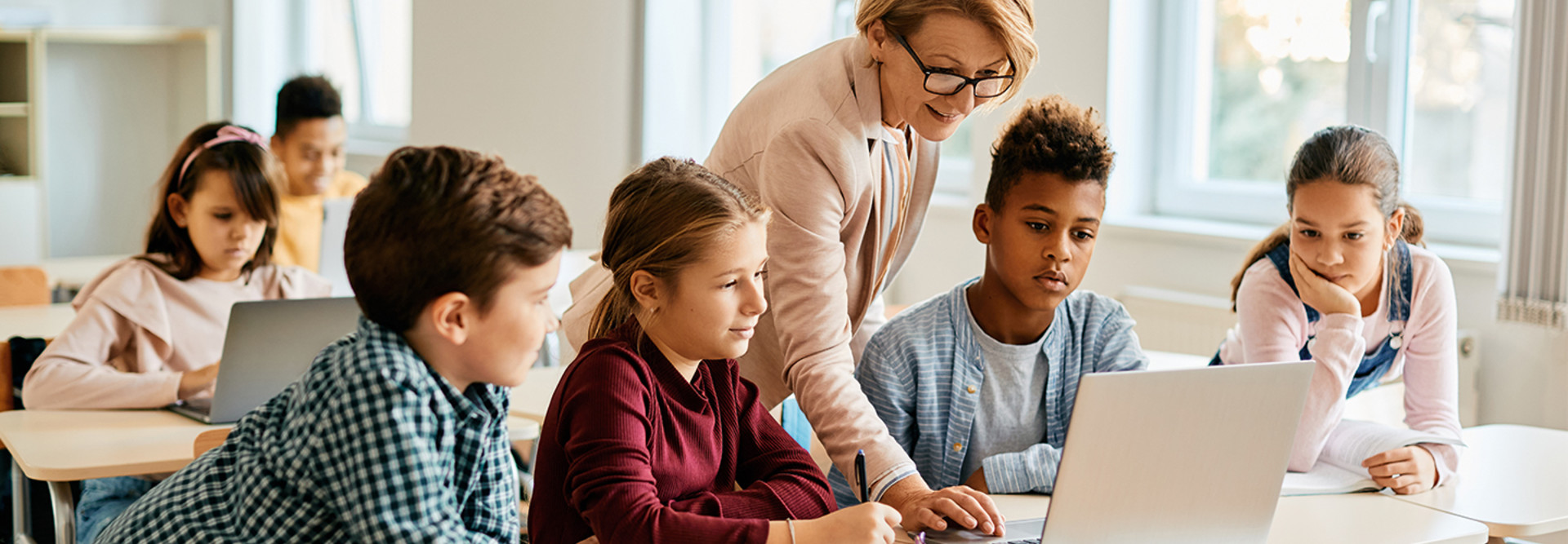 teacher helps student on laptop surrounded by other students