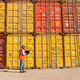 Supply Chain Disruptions - dock worker with laptop standing in front of cargo containers stacked two high