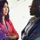 Image shows two women looking at each other with their arms crossed and a cityscape behind them