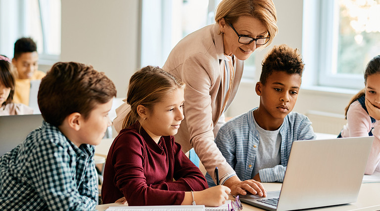 teacher helps student on laptop surrounded by other students