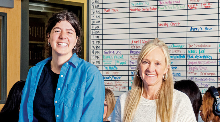 People in a classroom in front of a radio station scheduling board